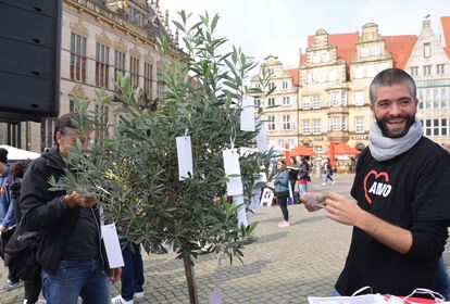 Lachender Mann trägt T-Shirt mit AWO-Logo vor ihm ein Olivenbaum in den Zettel gehängt sind bei einer Veranstaltung auf dem Bremer Marktplatz.
