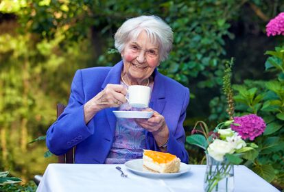 Lächelnde ältere Dame sitzt im Grünen an einem Tisch und trinkt Kaffee, vor sich ein Stück Kuchen.