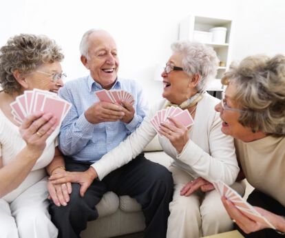 drei ältere Frauen und ein älterer Mann spielen gemeinsam Karten. Die Stimmung wirkt gelöst, sie lachen zusammen.