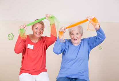 Pflegekraft und ältere Frau machen gemeinsam gymnastische Übungen. Sie halten beide ein buntes Tuch über dem Kopf
