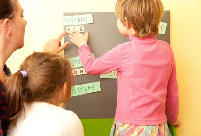 Ansicht von hinten: Kinder und Mitarbeitende formen das Wort Fisch mit Buchstaben auf einer magnetischen Tafel.