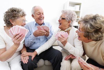 drei ältere Frauen und ein älterer Mann spielen gemeinsam Karten. Die Stimmung wirkt gelöst, sie lachen zusammen.